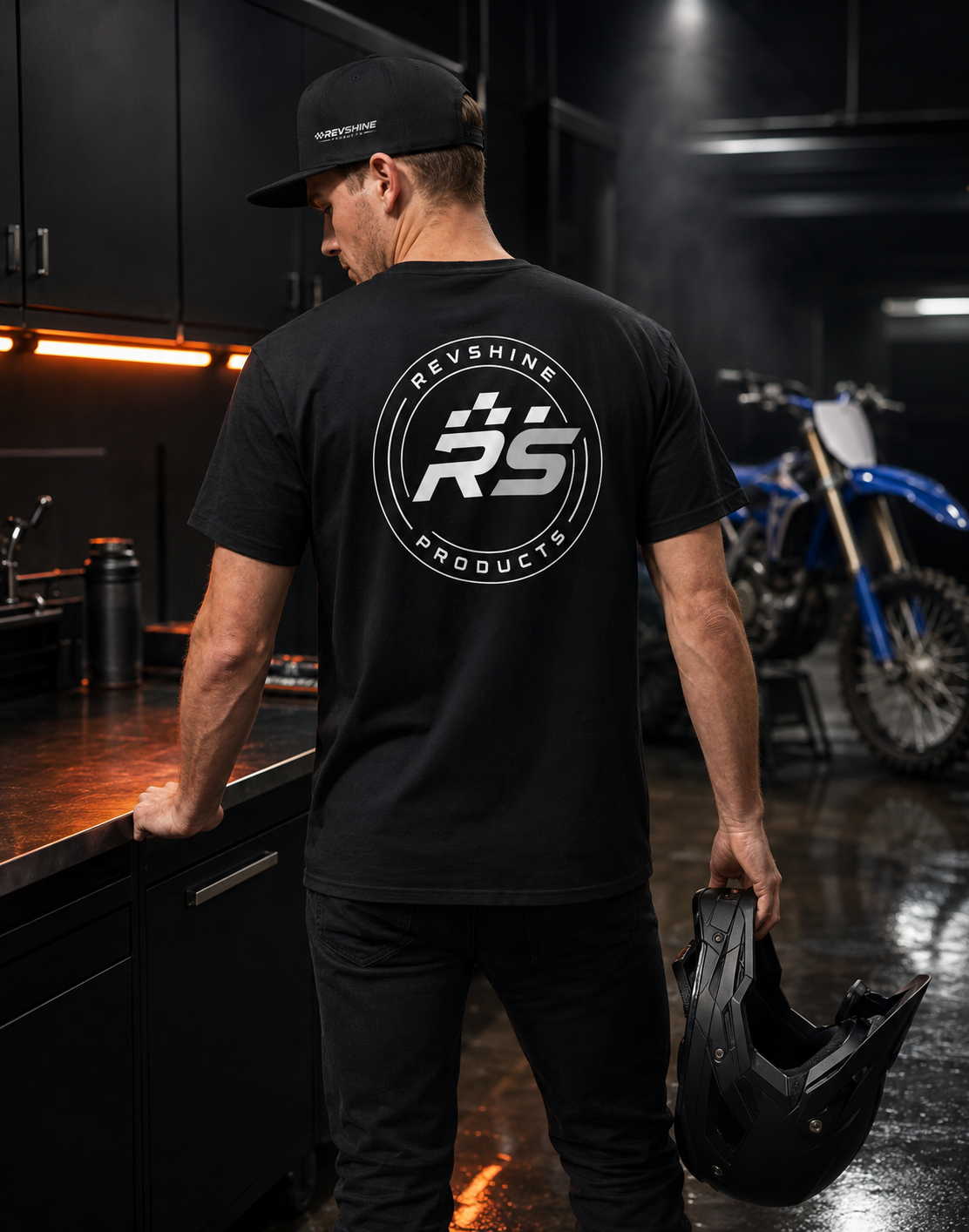 Rider wearing a black RevShine T-shirt and snapback while holding a motocross helmet in a premium workshop, with a blue dirt bike in the background.