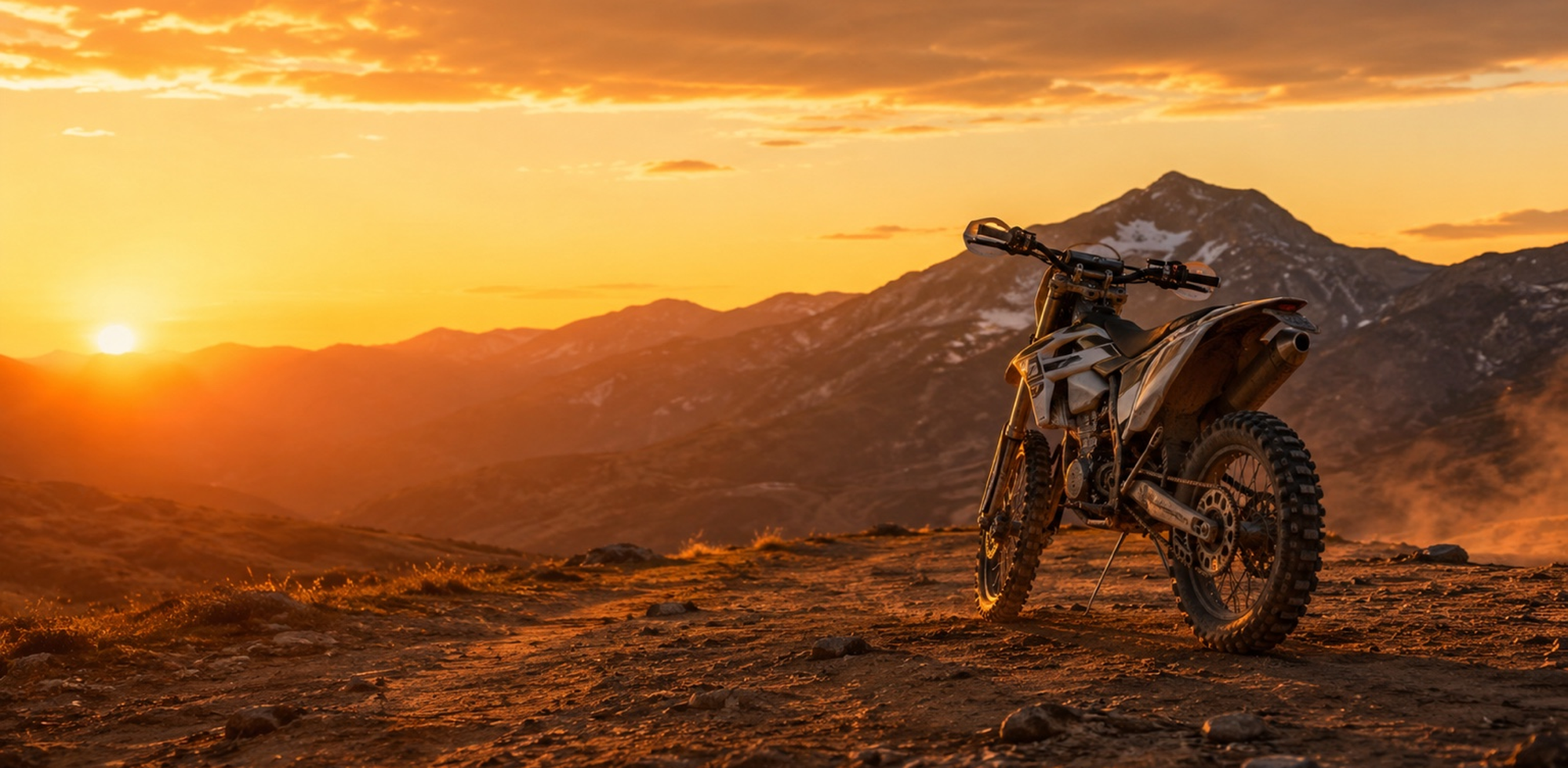 An off-road dirt bike is parked on a rocky mountain ridge at sunset, with dramatic orange and yellow clouds, a low sun, and dark mountain peaks in the background.