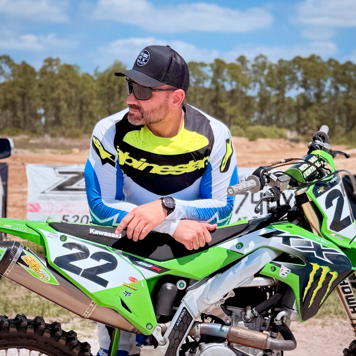 A man in a blue, white, and yellow Alpinestars motocross jersey leans on a green and white Kawasaki dirt bike, with trees and a blue sky in the background.
