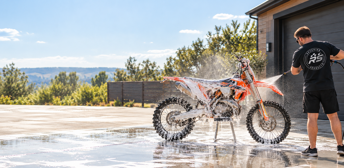 Motocross dirt bike covered in thick white cleaning foam being pressure-washed outdoors by a person wearing a black RevShine Products shirt. Premium off-road motorcycle cleaning in action.