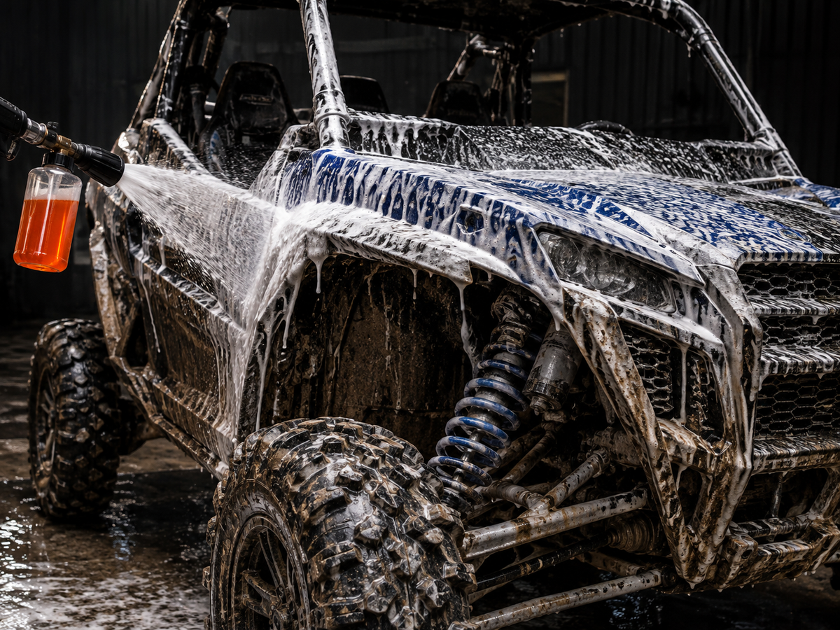 A muddy off-road vehicle covered in soapy foam while water sprays across it, with an orange liquid container held in a hose on the left.